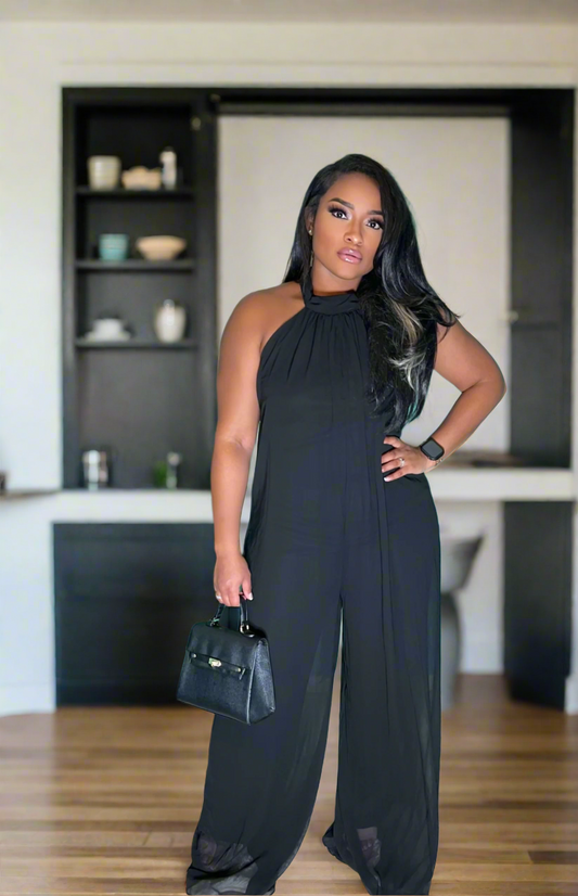 A woman standing in a backless black jumpsuit, posing with one hand on her hip. The jumpsuit is stretchy and has a high waist, with a V-neckline.